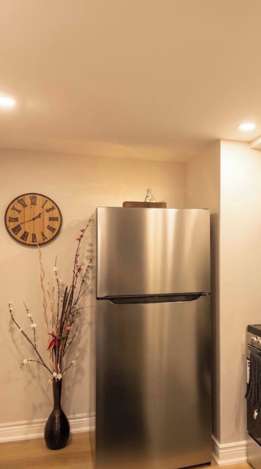 A stainless steel refrigerator in a corner of a room with a wall clock and a vase with branches.