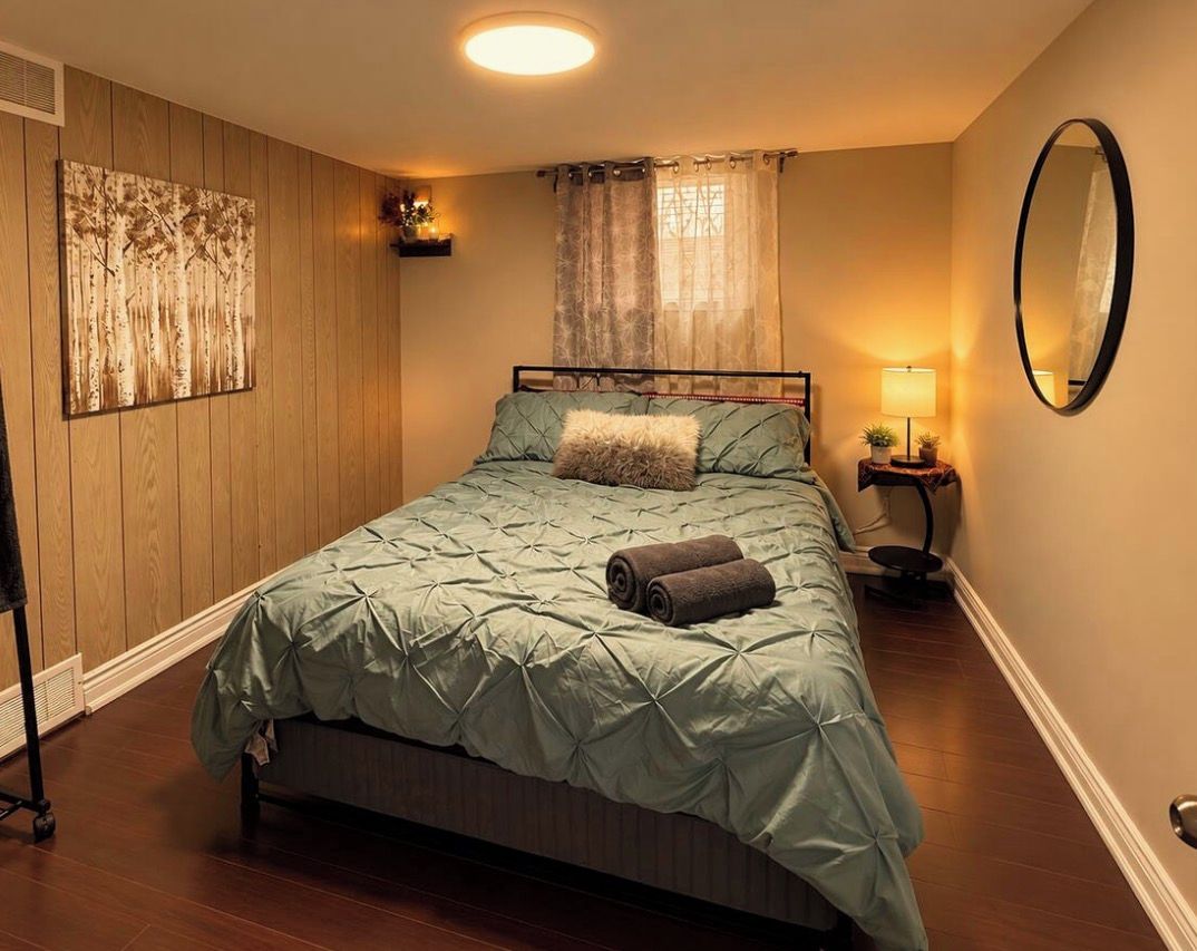 A bedroom with a bed, a bedside table with a lamp, a wall-mounted mirror, and a window with curtains.