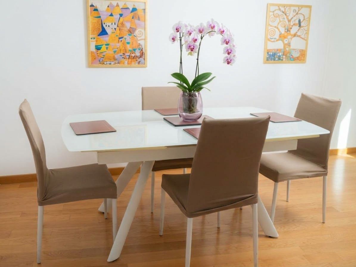 Zona pranzo con un tavolo bianco, quattro sedie marroni e un vaso con fiori.