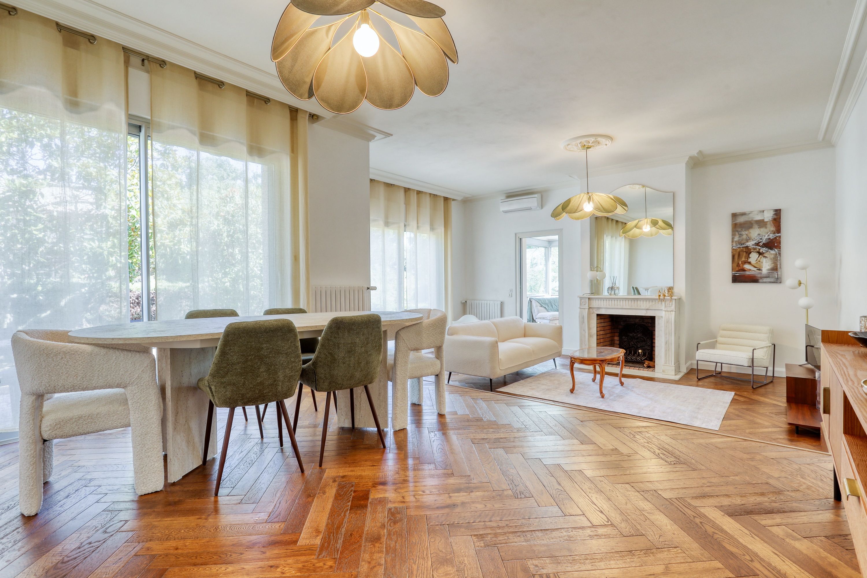 A spacious living room with a fireplace, a dining area with a round table and chairs, and a large window with curtains.