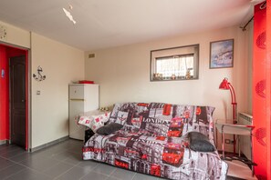 A living room with a sofa, a small table, a red door, a white refrigerator, and a window with curtains. - Clin d’Œil sur la Mer - 300m de la Plage (Cucq)
