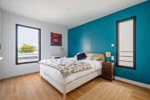 A bedroom with a large bed, a nightstand, a lamp, a painting, and a window with a view. - Ker Valy - Piscine privée et chauffée (La Trinite-sur-Mer)