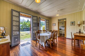 A dining area with a wooden table and chairs, a kitchen in the background, and a view of palm trees through open doors. - La Case Hoarau Duportail - Case créole du village (Entre-Deux)