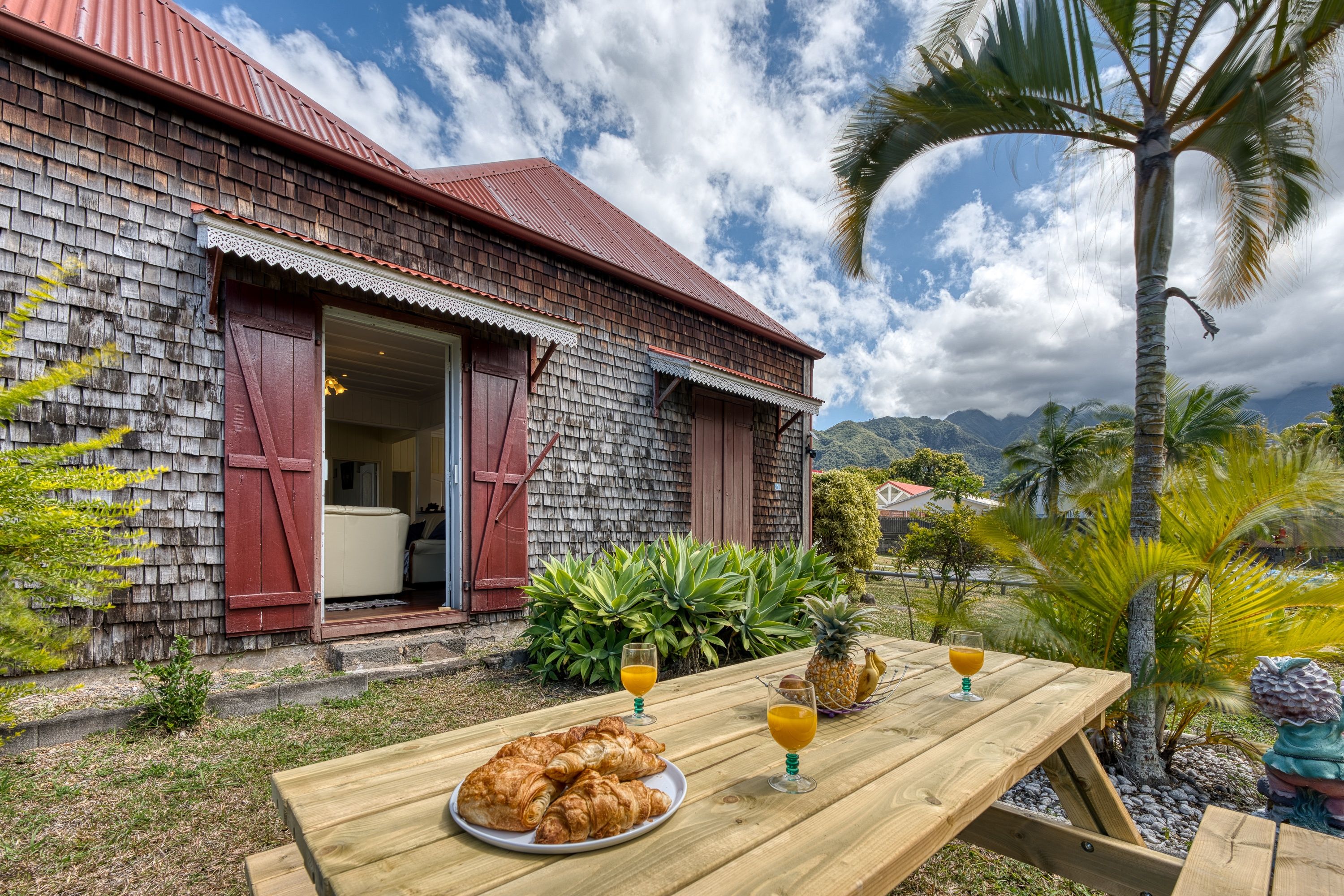 Une maison de campagne en bois, avec un toit rouge, une table à manger où sont disposés des croissants et des boissons, et une vue sur les montagnes et les palmiers.
