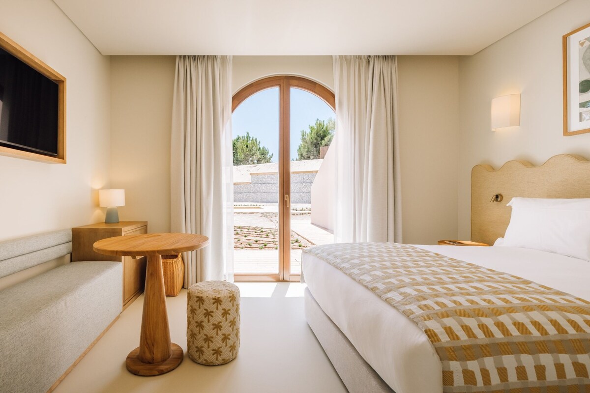 A hotel room with a bed, a small table, a lamp, a decorative stool, and a view through a glass door.
