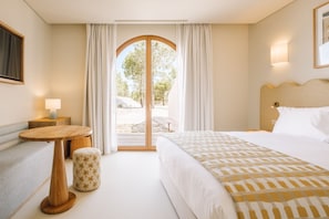 A hotel room with a bed, a small table, a chair, a lamp, and a view through a glass door. - Ando Living - Comporta Villas (Grândola)