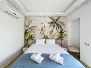 Ein Hotelzimmer mit einem großen Bett, zwei weißen Kissen, einer blauen Bettdecke und einem Wandgemälde mit tropischem Motiv.