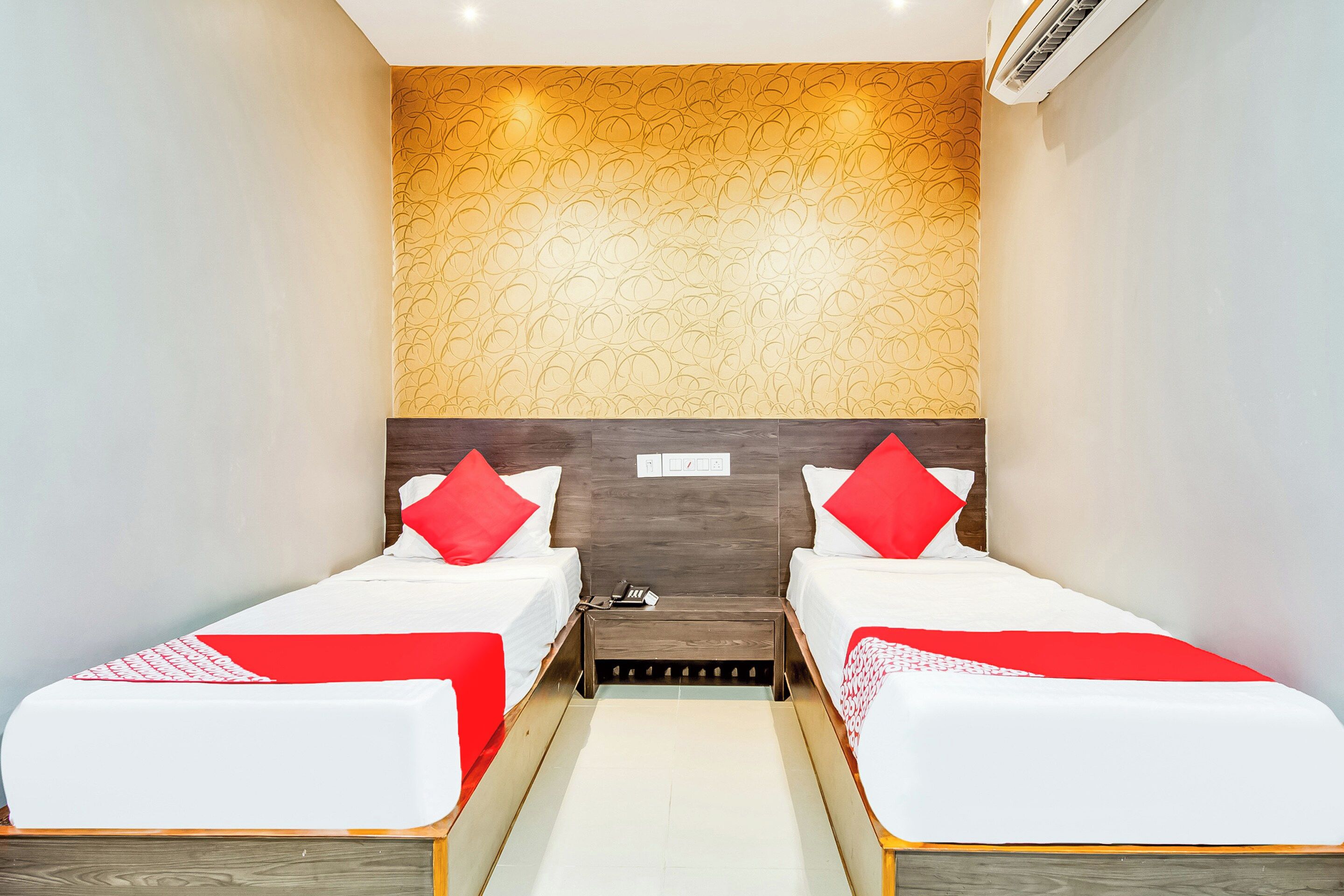 A hotel room with two single beds, a wooden headboard, a wall-mounted air conditioner, and a small bedside table with a phone.