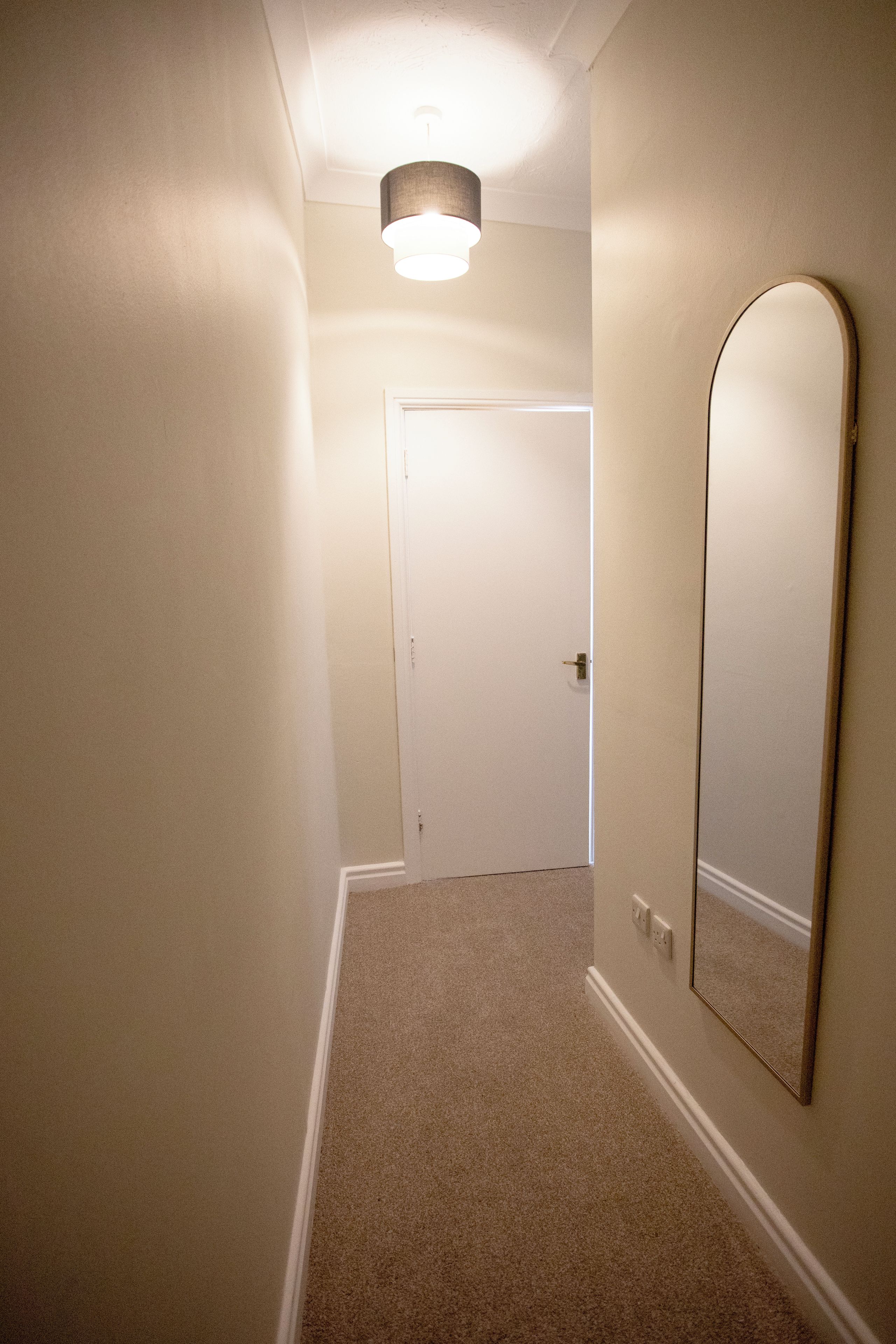 A narrow hallway with a carpeted floor, a door at the end, and a wall-mounted mirror.