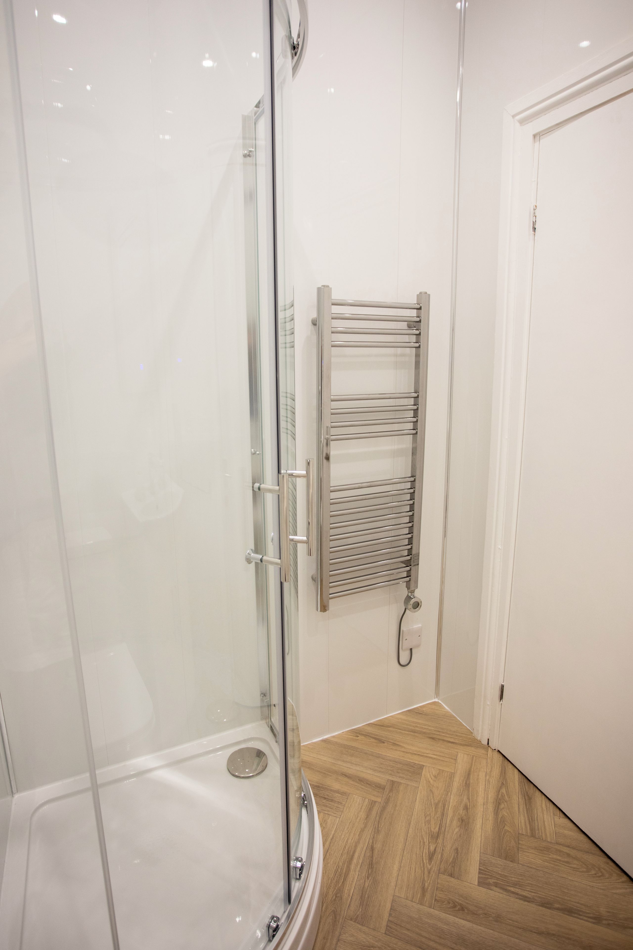 A glass-enclosed shower with a white tiled wall and a wooden floor.