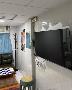 Habitación con una televisión de pantalla plana grande montada en la pared, un sofá, una pequeña mesa con mantel a cuadros y un taburete.
