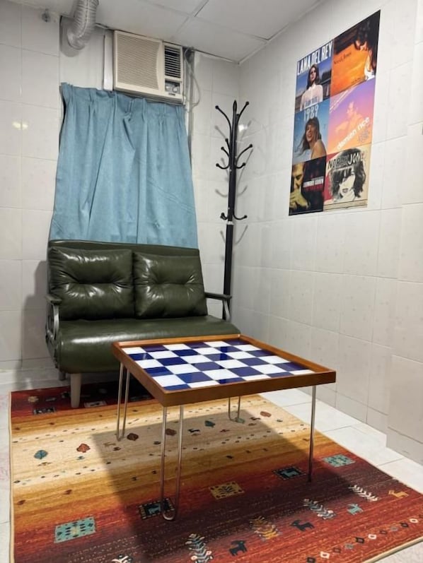 Una habitación con un sofá de cuero verde, una pequeña mesa de madera con superficie a cuadros, una alfombra estampada y una pared con carteles.