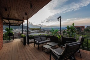 Eine Holzterrasse mit Gartenmöbeln, darunter eine Couch und Stühle, mit Blick auf eine Stadtlandschaft mit Gebäuden und Bergen.