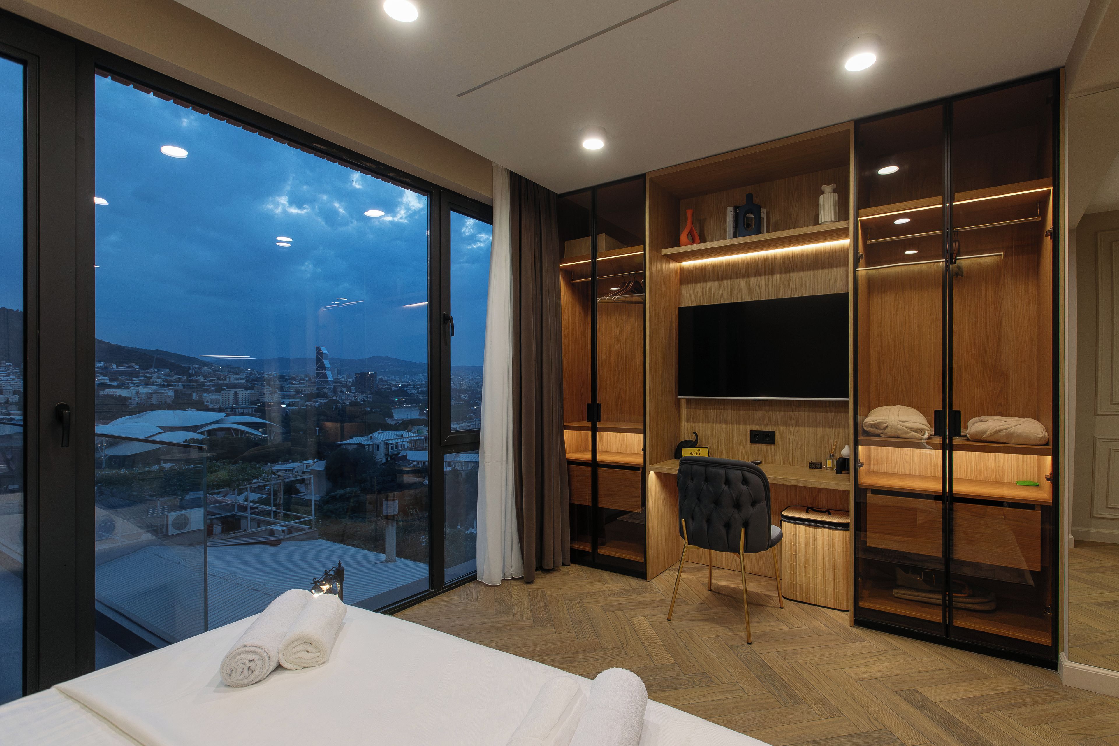 A modern hotel room with a large sliding glass door, a bed with white linens, a seating area with a chair, and a built-in TV shelf.