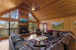 A spacious living room with a fireplace, leather sofas, a television, and large windows offering a view of the outdoors. - Family Friendly-Game Room-Hot Tub-Fireplace (Sevierville)