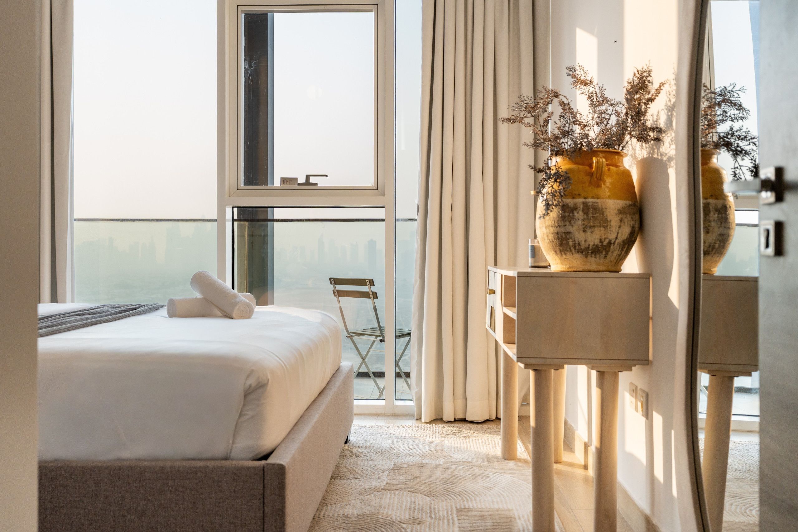Une chambre d’hôtel avec un lit, une chaise, une table avec un vase et une vue sur la ville par la fenêtre.