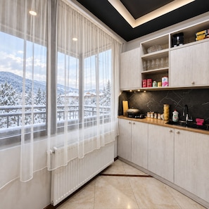 A modern kitchen with a view of snow-covered mountains, a window with sheer curtains, and a black countertop with various kitchen items.