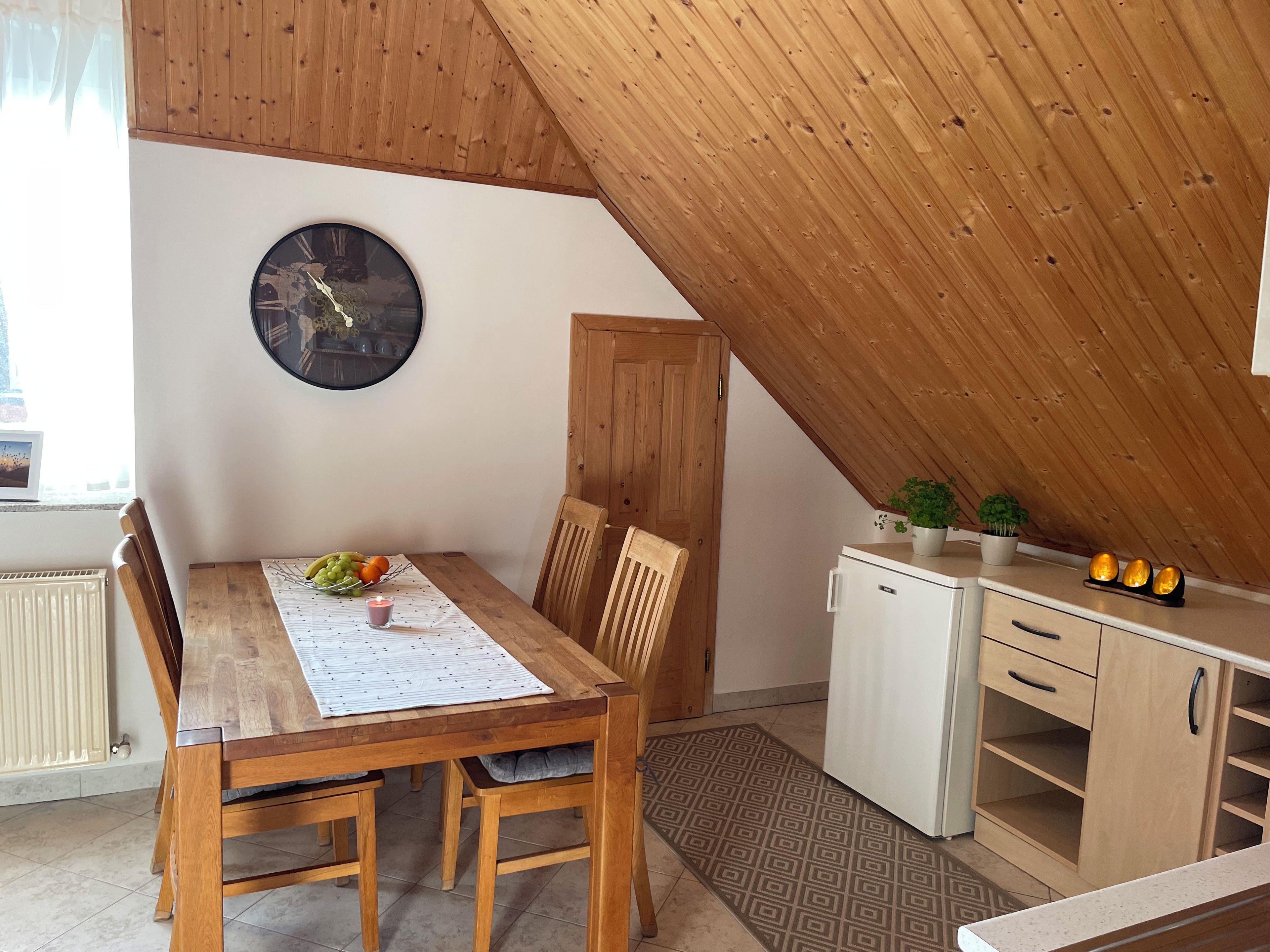 A dining area with a wooden table, chairs, a clock, and a small refrigerator.