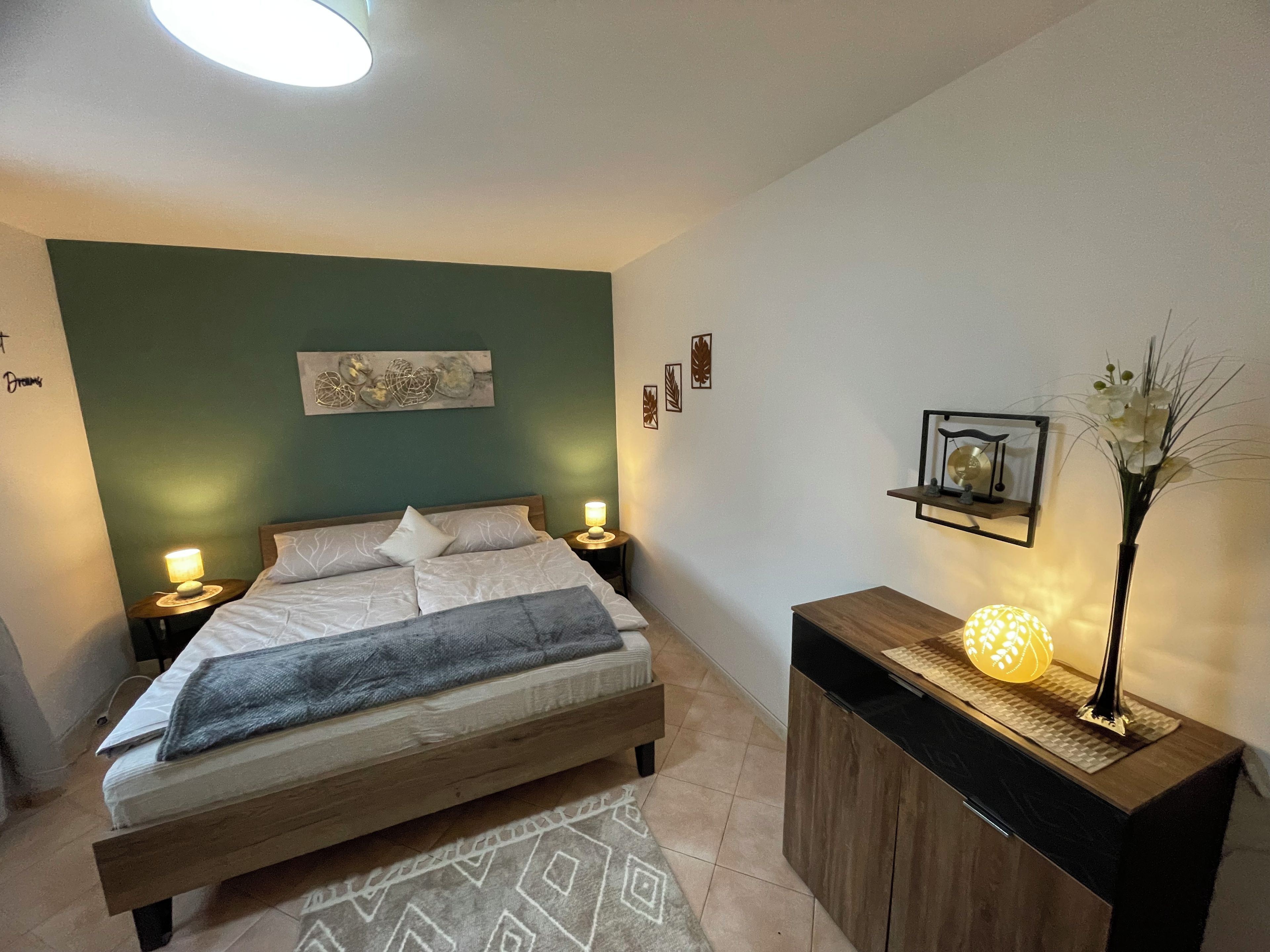 Une chambre à coucher comprenant un lit, des lampes de chevet, une table de chevet, un élément décoratif mural, une petite étagère avec une lampe et un vase avec des fleurs.
