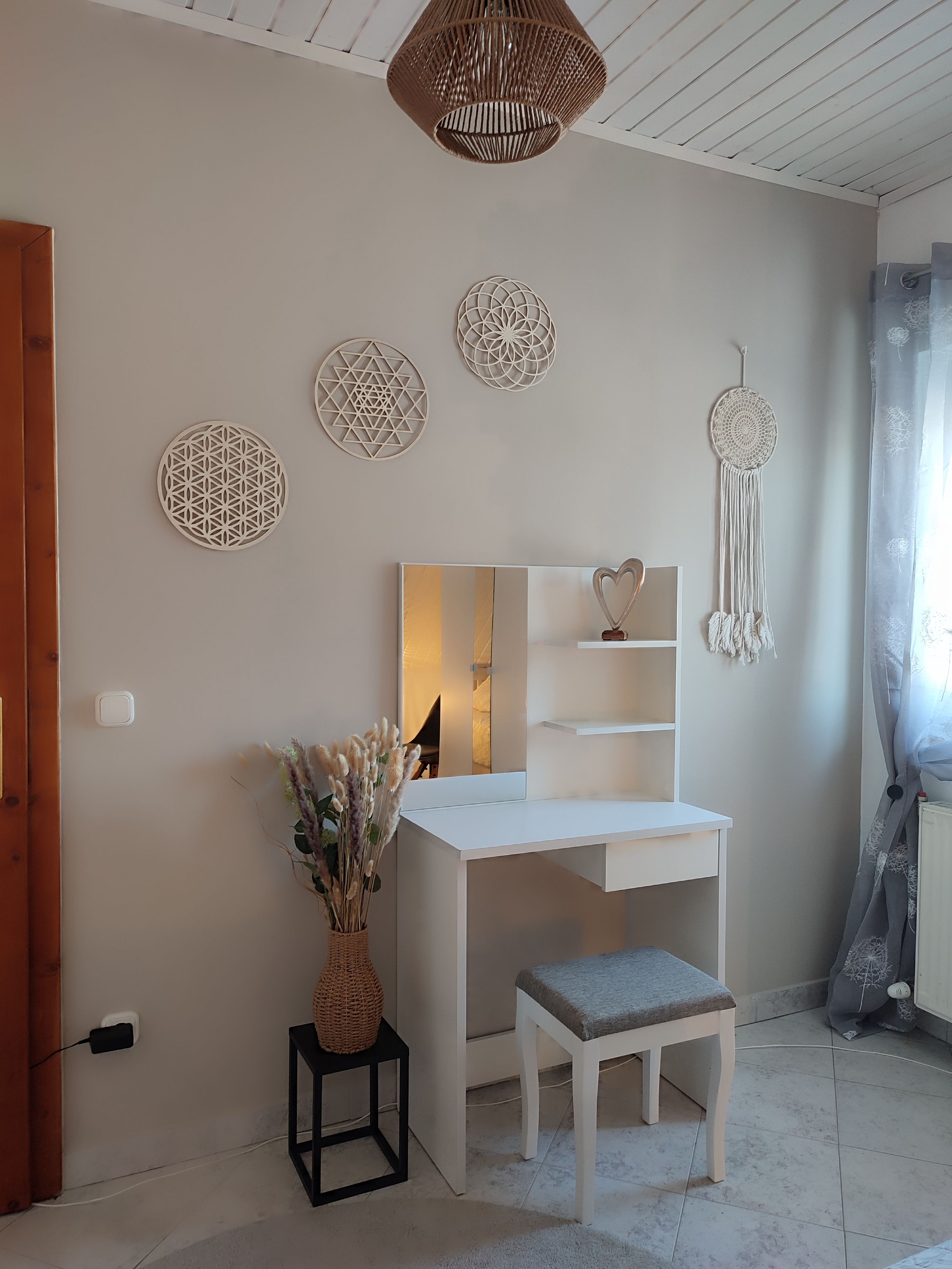 Une chambre moderne avec un bureau blanc, un miroir, des œuvres d’art décoratives au mur et un attrape-rêves suspendu.