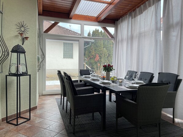 A dining area with a table set for four, surrounded by wicker chairs, a decorative lantern, and a view of a house and trees through a glass door.
