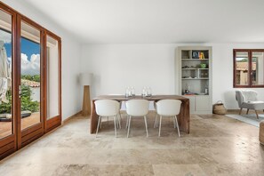 Un comedor con una mesa de madera y cuatro sillas, una estantería con libros y objetos decorativos, y vistas a un jardín a través de amplios ventanales.