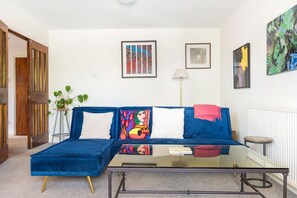 A modern living room with a blue sofa, a glass coffee table, and a potted plant. - GuestReady - Charming North London Retreat (London)