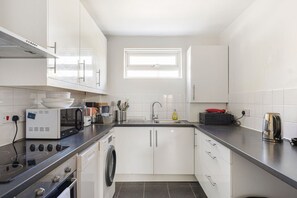 A modern kitchen with white cabinets, a built-in oven, a washing machine, and a window above the sink. - GuestReady - Charming North London Retreat (London)