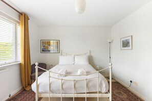 Ein Bett mit weißem Gestell und weißer Bettwäsche in einem Zimmer mit Fenster, Stehlampe und zwei gerahmten Bildern an den Wänden.