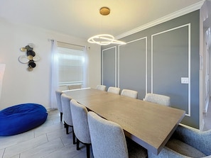 A modern dining area with a long table, grey upholstered chairs, a blue bean bag, and a unique circular light fixture. - Sunshine Retreat: Private Pool and Spa! (Davenport)