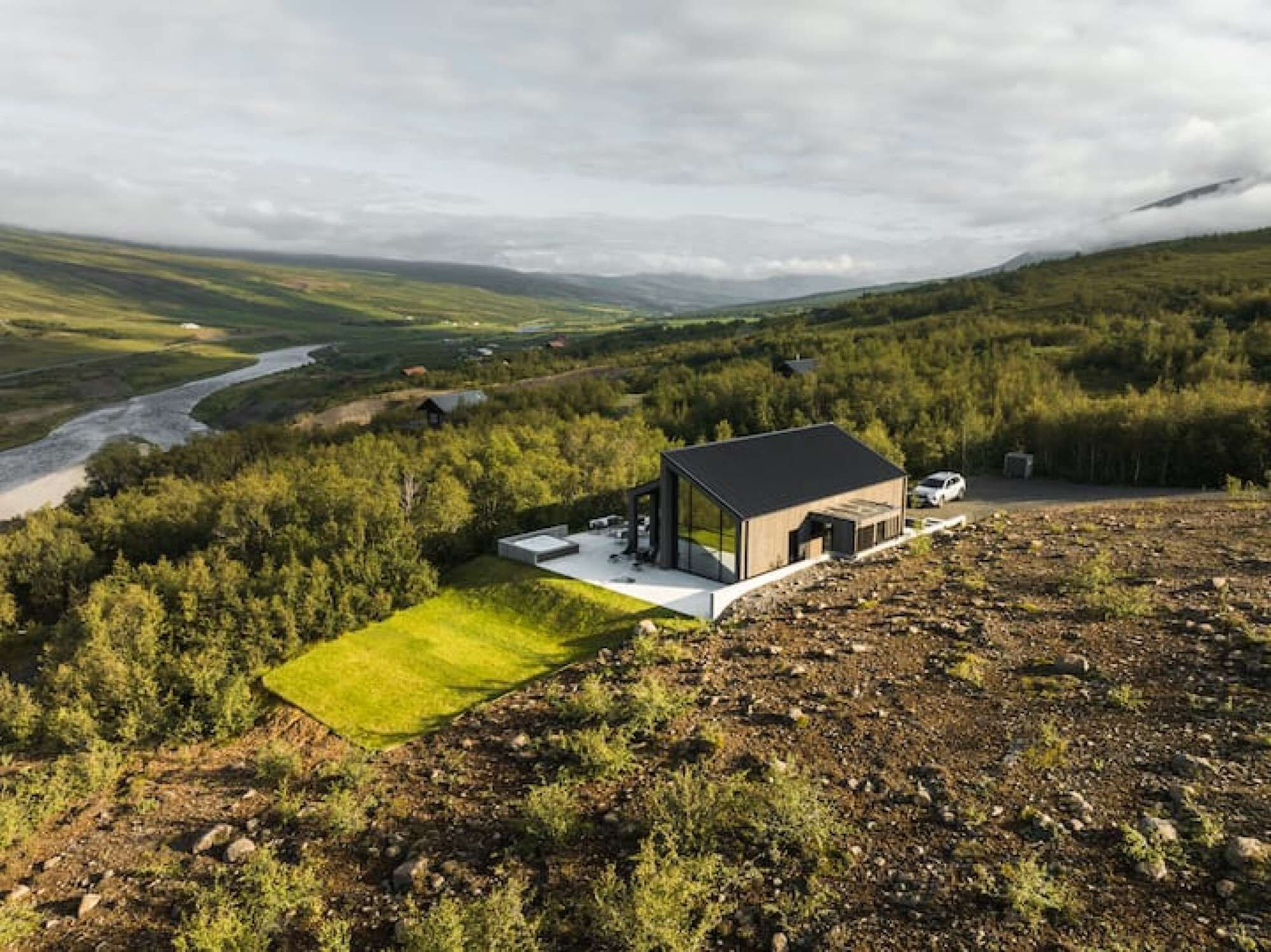 A modern house with a glass facade, a paved patio, and a view of a river and forested hills.