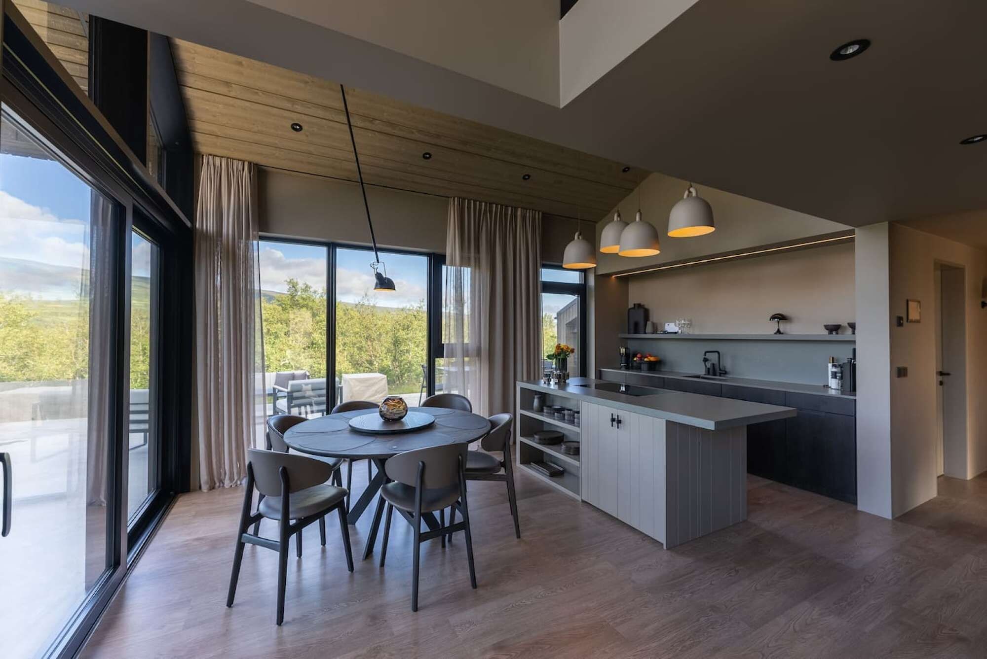 Een moderne keuken met een centraal eiland, een eetgedeelte met stoelen en grote ramen met uitzicht op de omgeving.