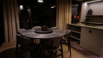 Un comedor con una mesa redonda y sillas, una zona de cocina con armarios y una encimera, y vistas a través de una ventana con cortinas.