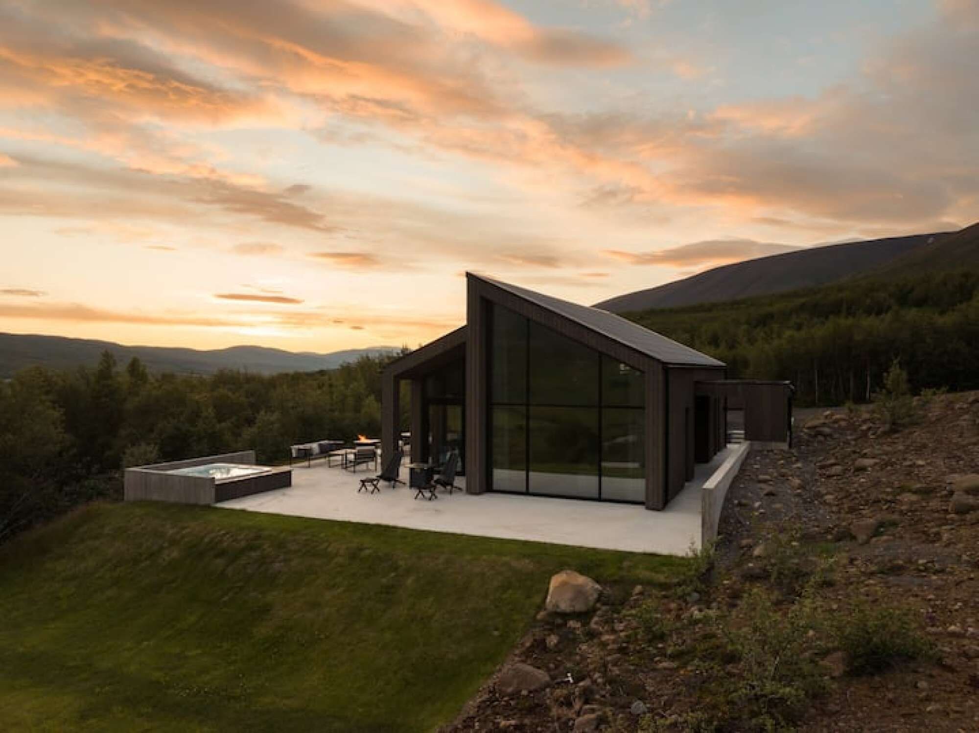 Una casa moderna con un amplio ventanal, un patio con mobiliario exterior y vistas al bosque y las colinas circundantes.