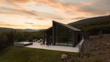 Una casa moderna con un amplio ventanal, un patio con mobiliario exterior y vistas al bosque y las colinas circundantes.