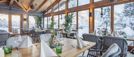 Un restaurante con vigas de madera, ventanas amplias y vistas a montañas nevadas.