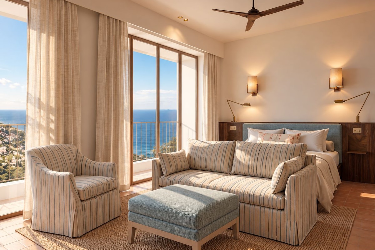 A hotel room with a large balcony, a bed with a headboard, two armchairs, a sofa, and a small ottoman.
