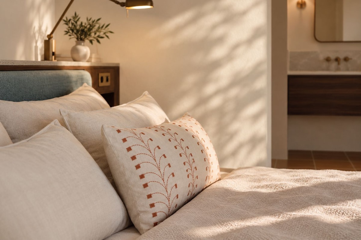 A bedroom with a bed, pillows, a bedside lamp, a nightstand with a plant, and a bathroom with a mirror and sink.