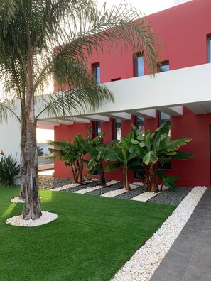 A modern building with a red facade, a covered outdoor area, and a well-maintained garden with palm trees and artificial grass. - Holiday Home 'Casa Da Brava' with Private Pool, Wi-Fi and Air Conditioning (Alvor)