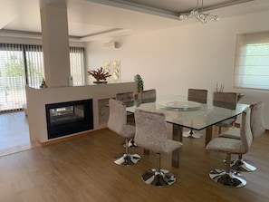 A modern dining area with a glass table and cushioned chairs, a fireplace, and a large window with blinds. - Holiday Home 'Casa Da Brava' with Private Pool, Wi-Fi and Air Conditioning (Alvor)
