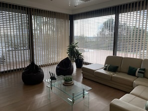 Ein modernes Wohnzimmer mit einem Glastisch, einem beigefarbenen Sofa und einem großen Fenster mit Jalousien.
