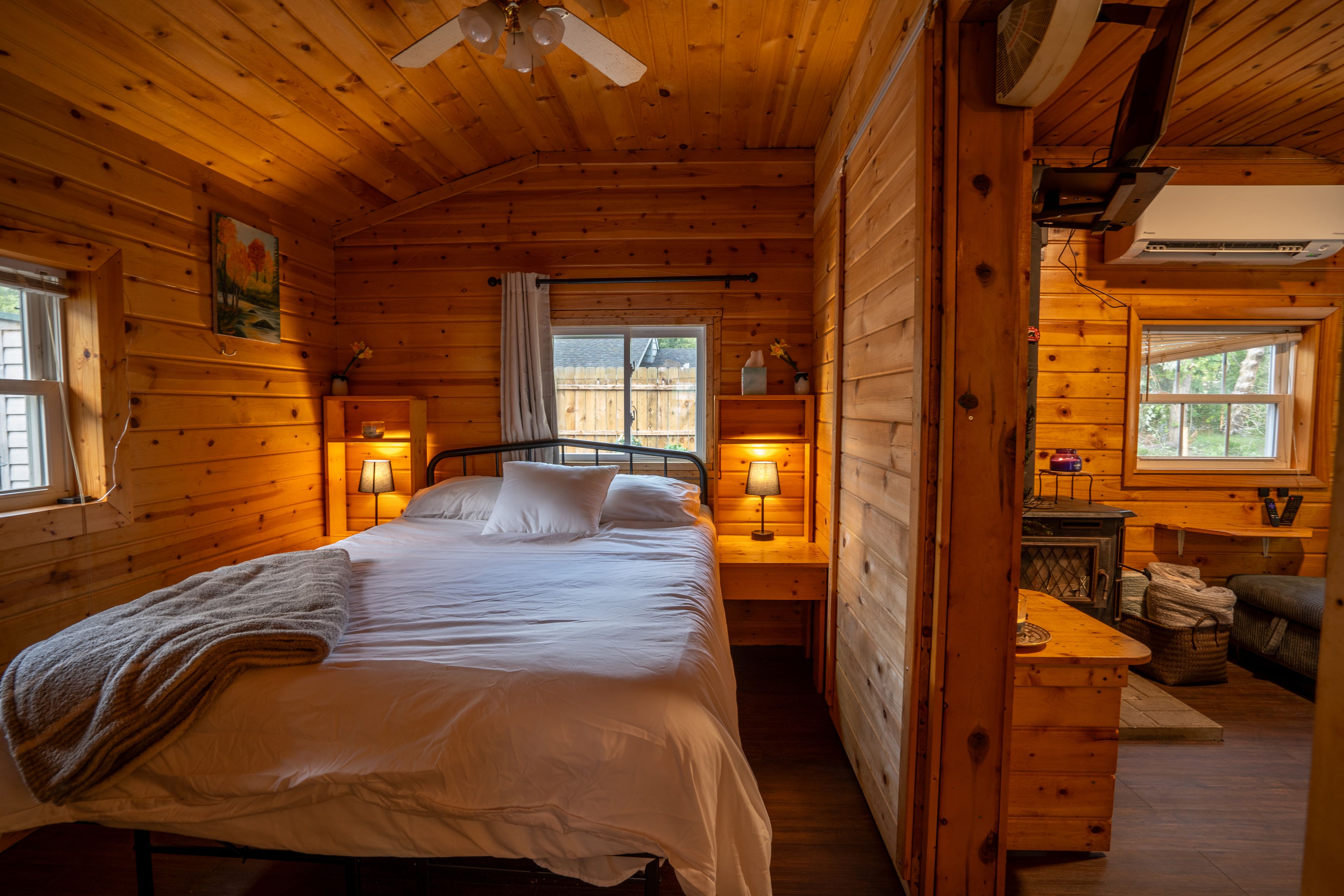 A wooden cabin room with a bed, bedside lamps, a window, and a small table with a chair.