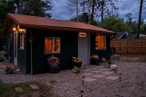 Ett litet, mörkgrönt hus med ett rött tak, en ytterdörr och två fönster. Framför huset står blomkrukor med blommor i.