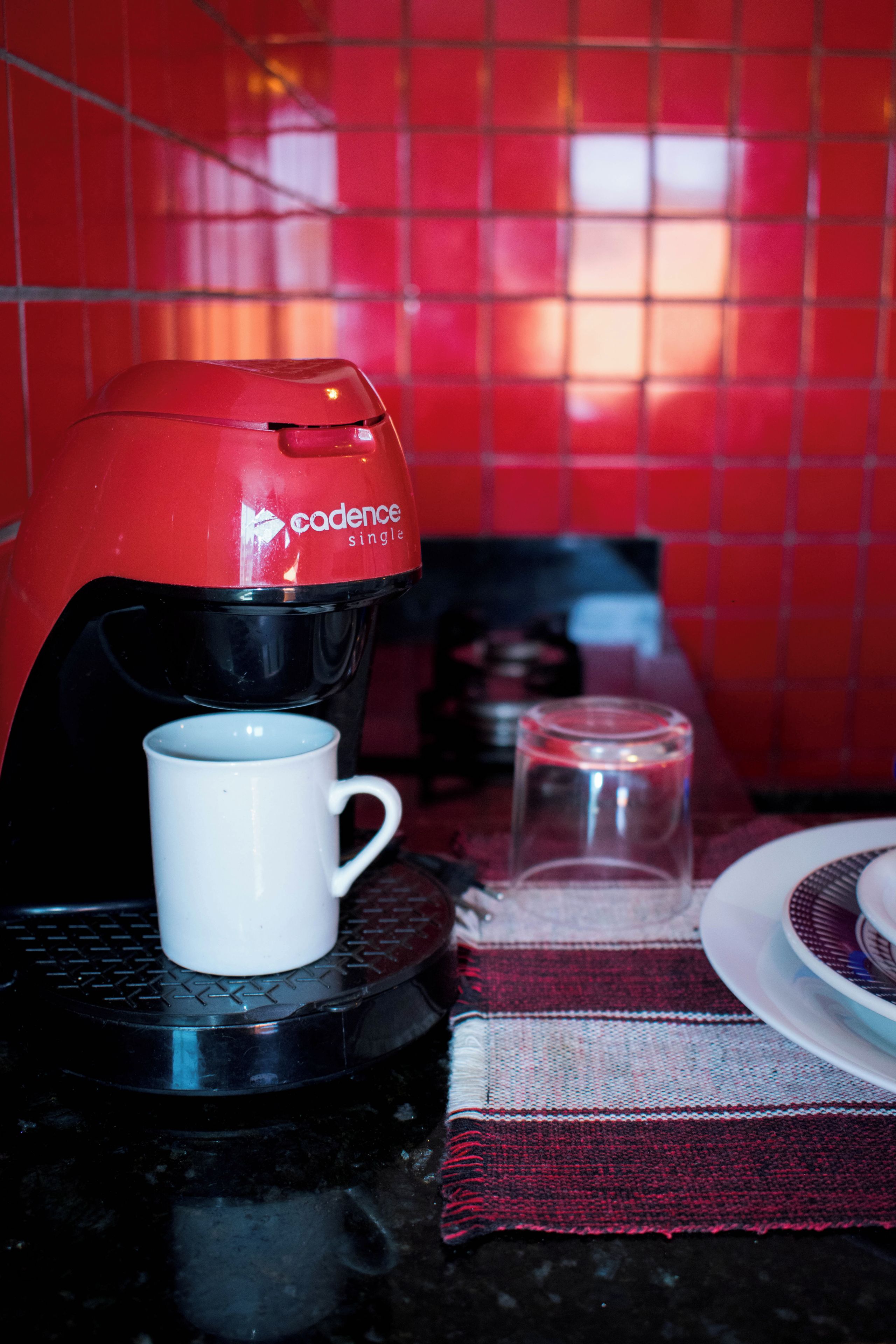 A coffee maker with a white mug on a black surface, a glass, and a plate with a cup on a red tiled wall.