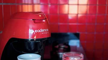 A coffee maker with a white mug on a black surface, a glass, and a plate with a cup on a red tiled wall.