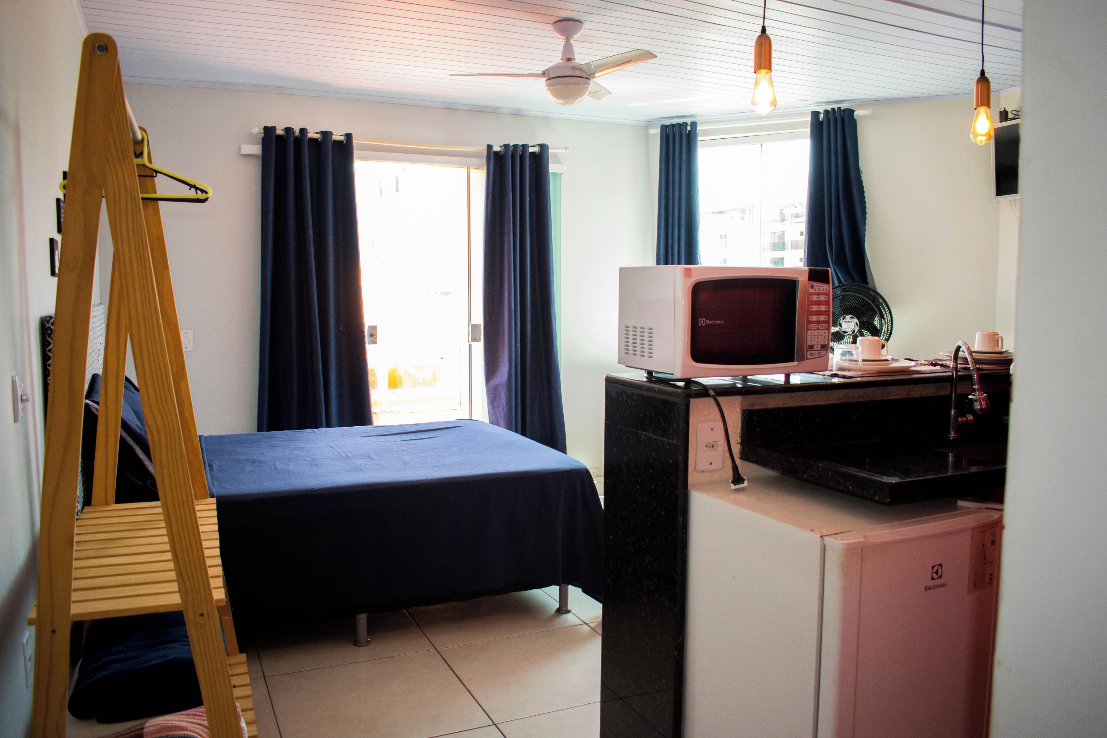 A compact hotel room with a bed, a desk with a microwave, a small refrigerator, a ceiling fan, and a window with curtains.
