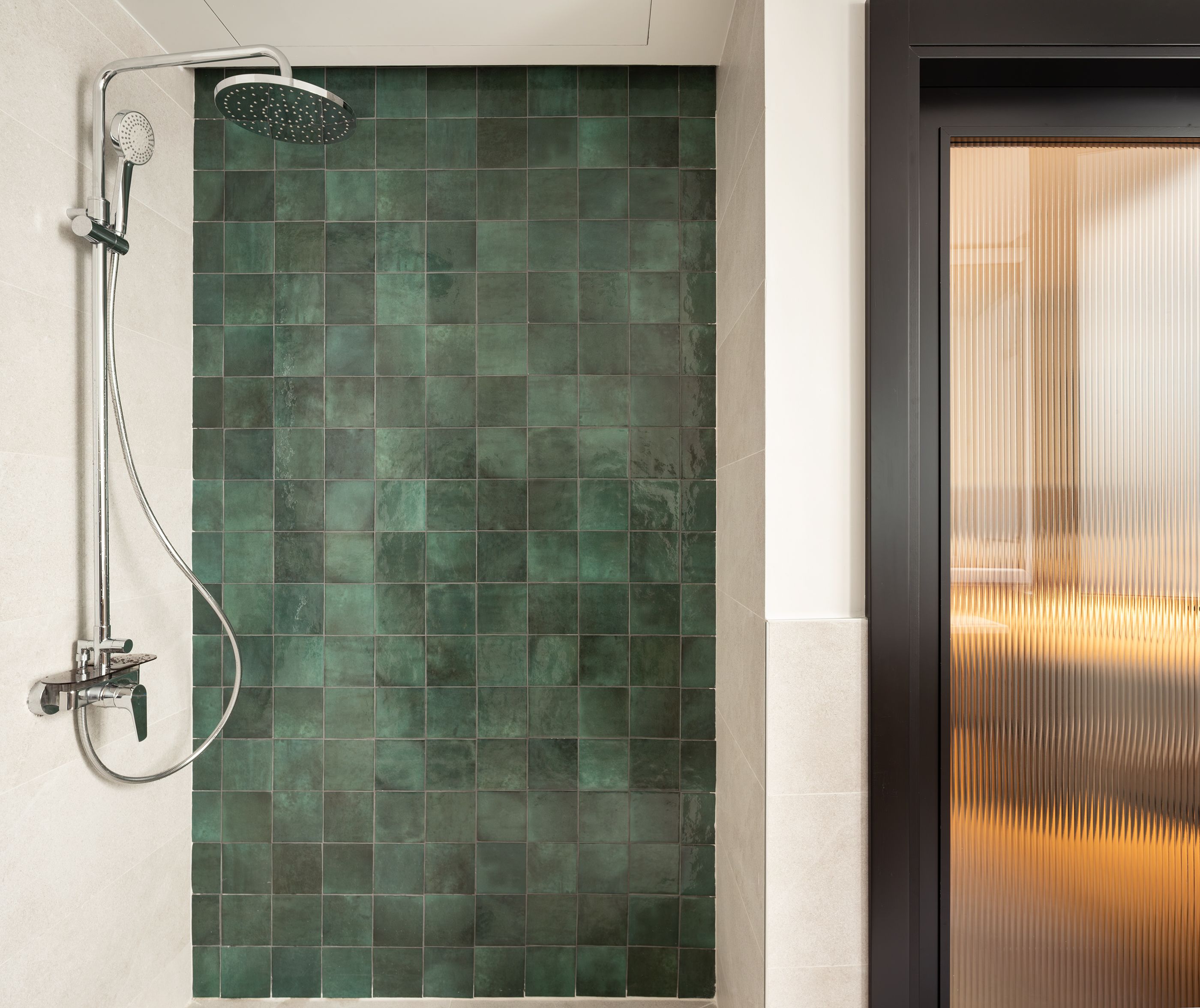 Een douche met een handdouche en een kraan op de muur, een betegelde muur met een groenachtige tint, en een deur met verticale zonwering.