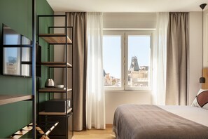 Habitación de hotel con cama, ventana con cortinas, televisor montado en la pared y estantería con varios objetos.