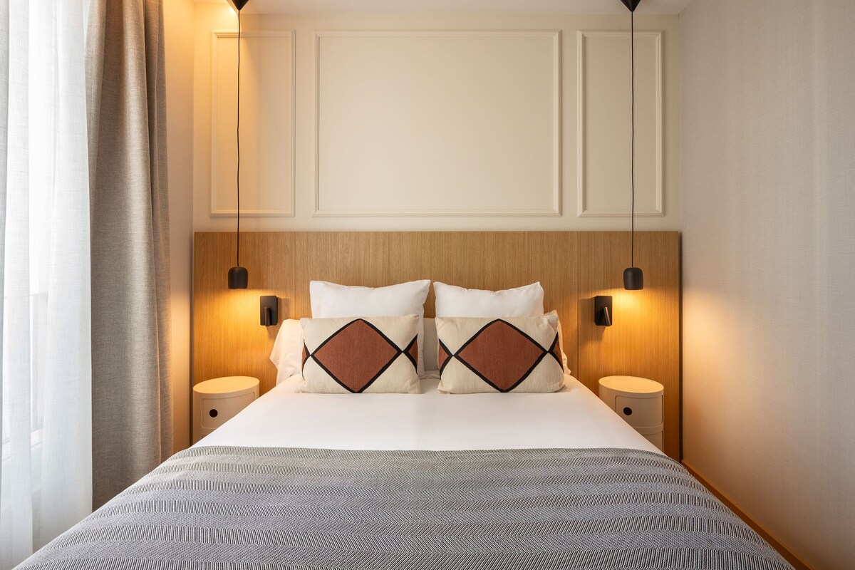 Una habitación de hotel con una cama, dos mesitas de noche, un cabecero y lámparas fijadas a la pared.