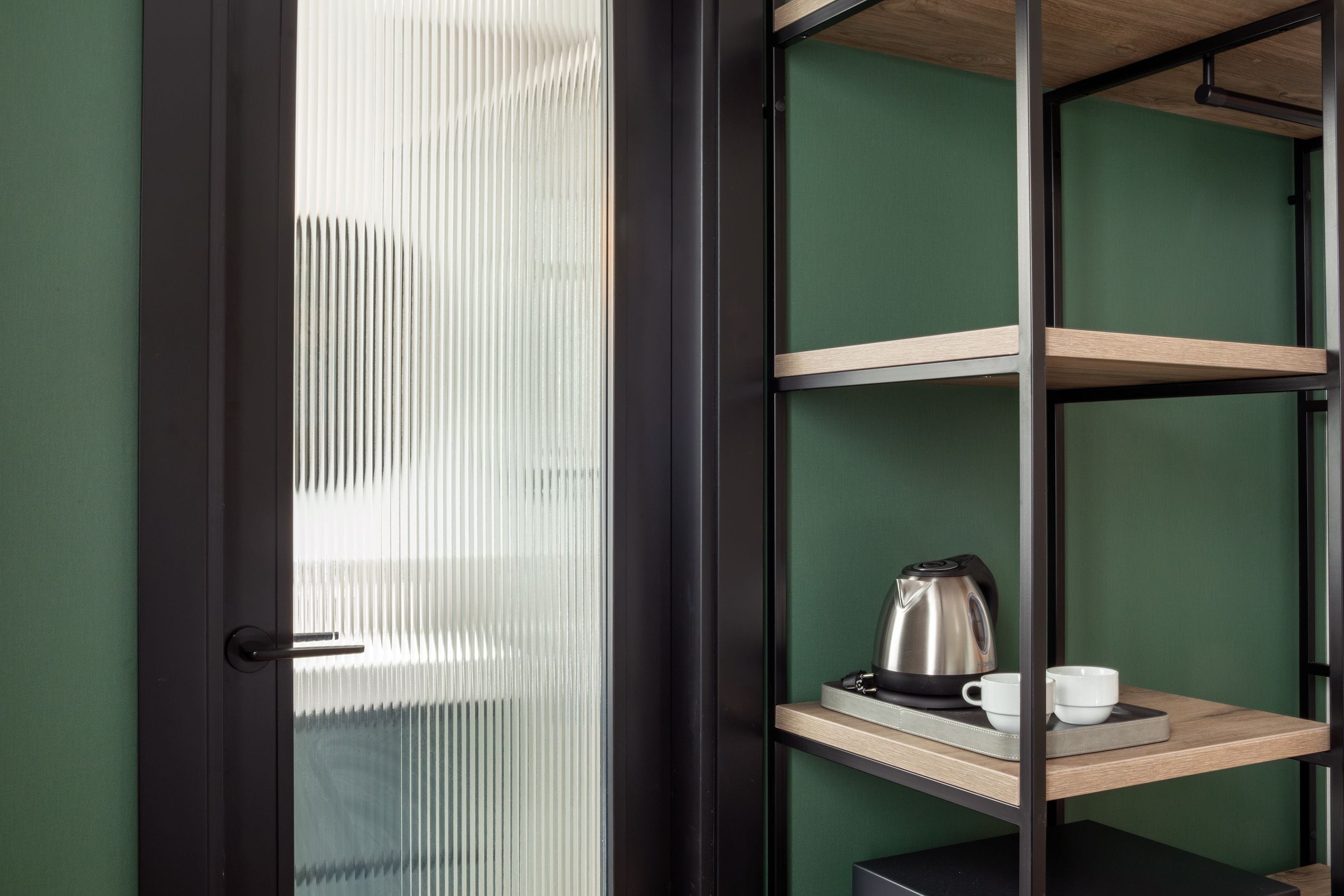 Een kamer met een deur, een plank met een waterkoker en kopjes, en een groene wand.